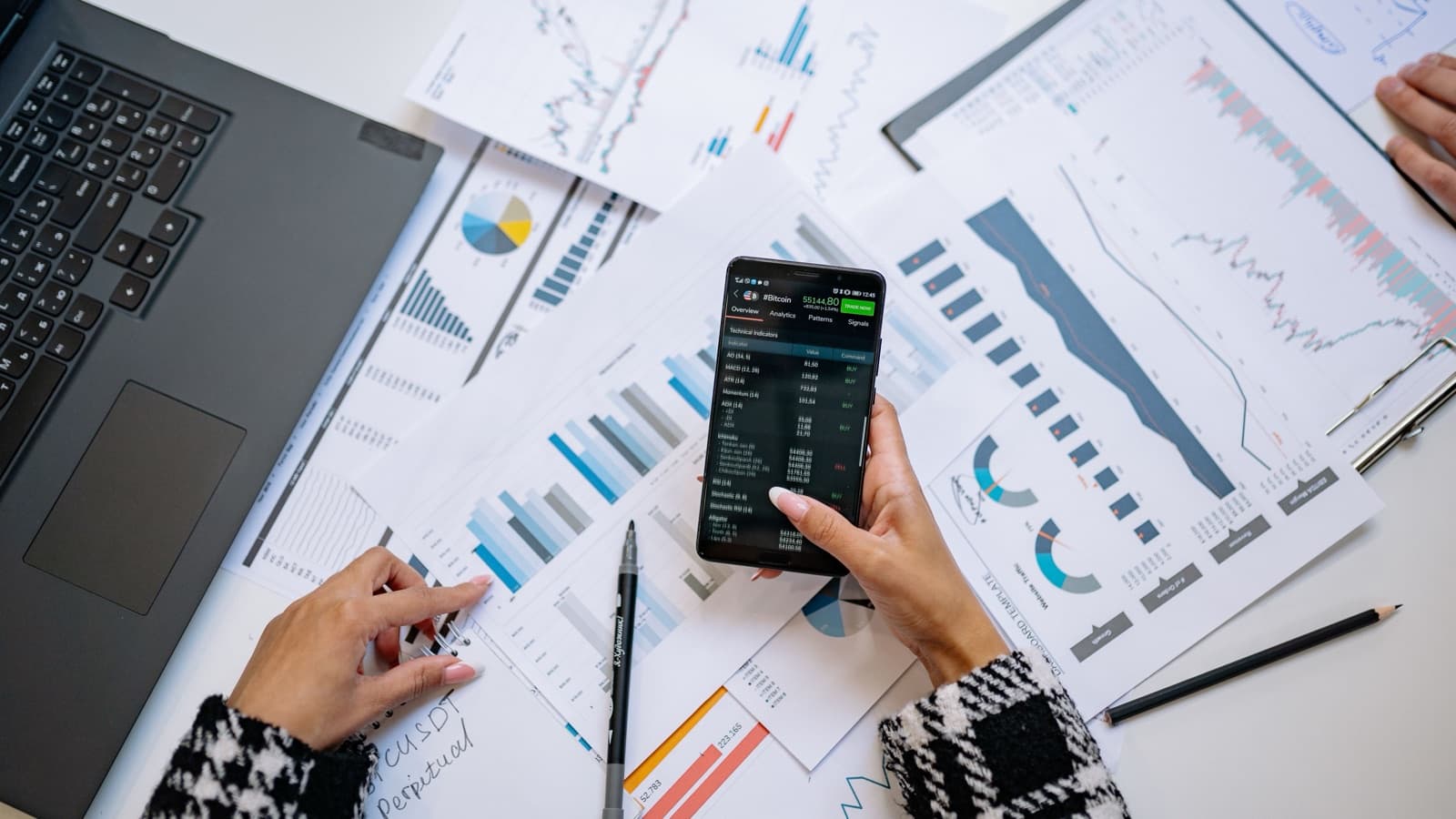 A person reviewing financial charts on a phone beside a laptop and printed reports